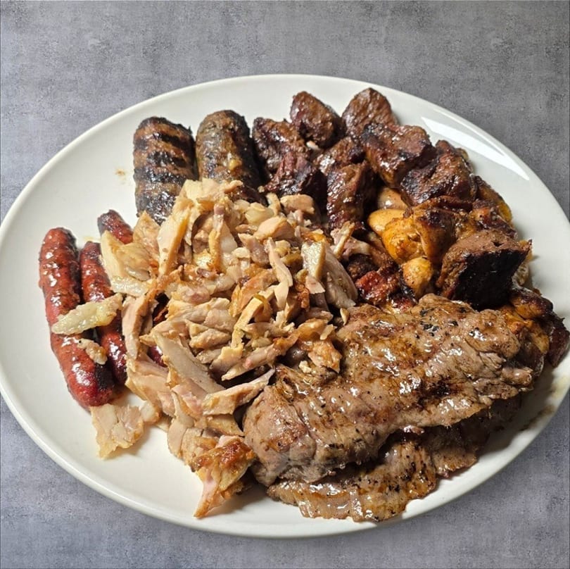 A white plate piled high with grilled meats, including sausages, shredded chicken, and steak.
