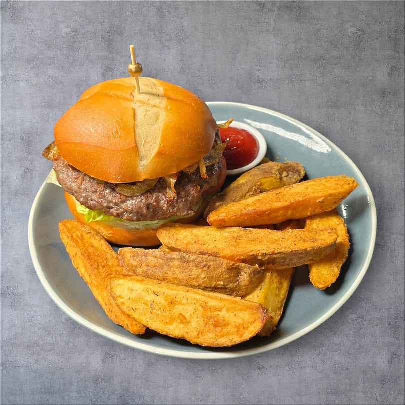 A beef burger with lettuce and caramelized onions, served with potato wedges and ketchup on a blue plate.