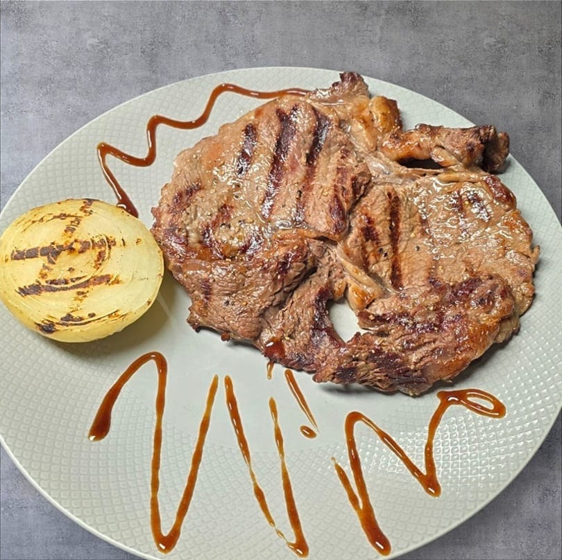 Grilled steak with grill marks and a grilled onion half, drizzled with sauce on a white plate.