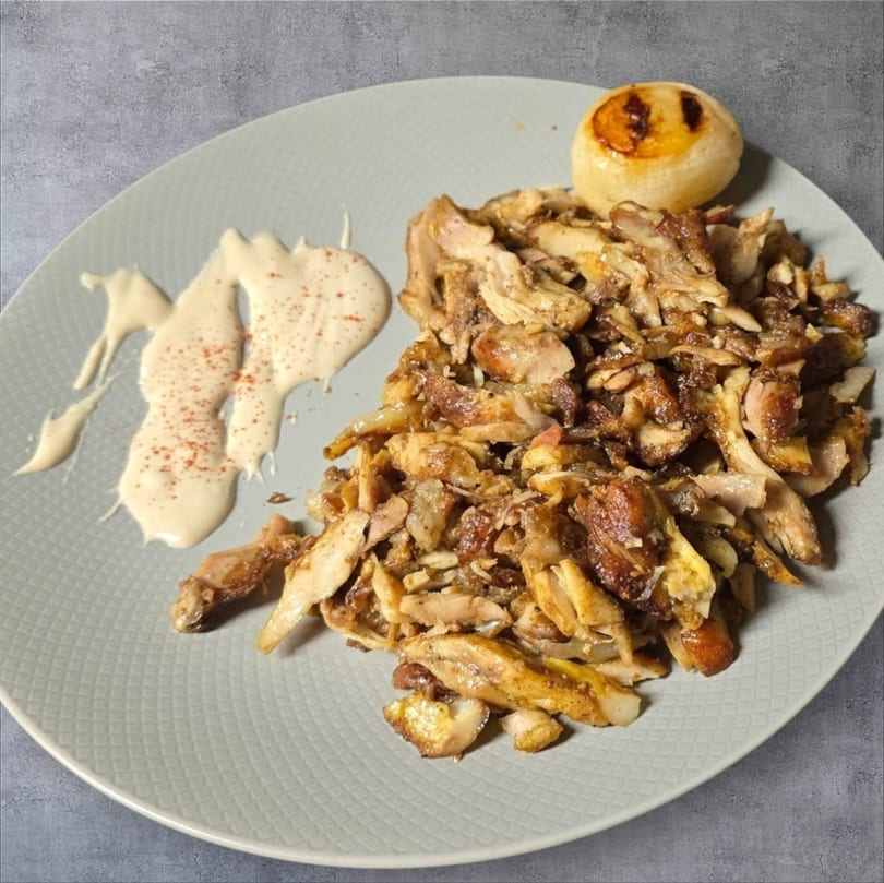 A plate of chicken shawarma with tahini sauce and a small bun on a gray plate.